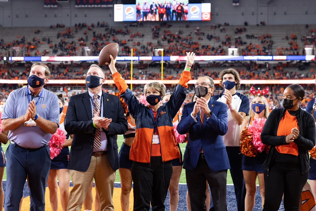 Eileen Collins stands on the ground floor of a football stadium with five others who are clapping. Collins is holding both arms high, and is holding a football.