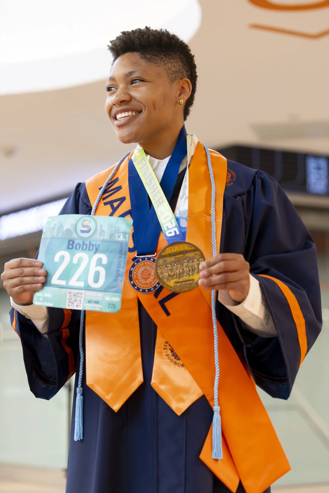 Bobby Marie Battle '26 standing in her graduation regalia and holding her half-marathon race bib and medal.