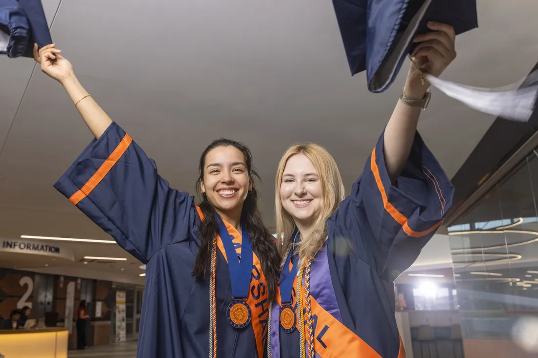 Student Marshals Ailed Torres Espinosa and Anna Meehan standing side by side holding up their graduation caps and smiling.