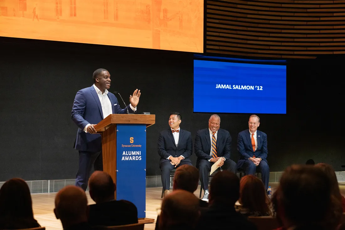 Wide shot of Salmon speaking at a podium in front of an audience, with other members on stage.