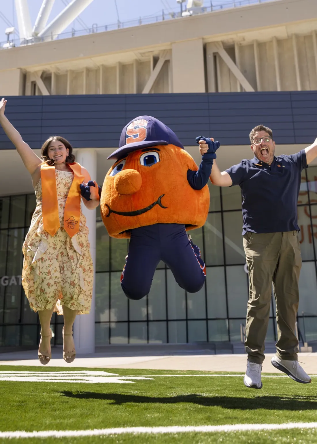 Sarah Jacoby '26 and Mitch Messinger ’92, G’93 holding hands with Otto the Orange and jumping in front of the JMA Wireless Dome.