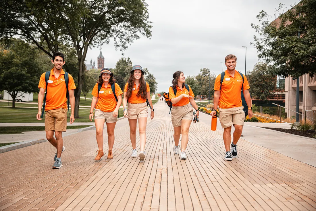 Five students wearing orange Orientation Leader tee shirts walk on the Einhorn Walkway.