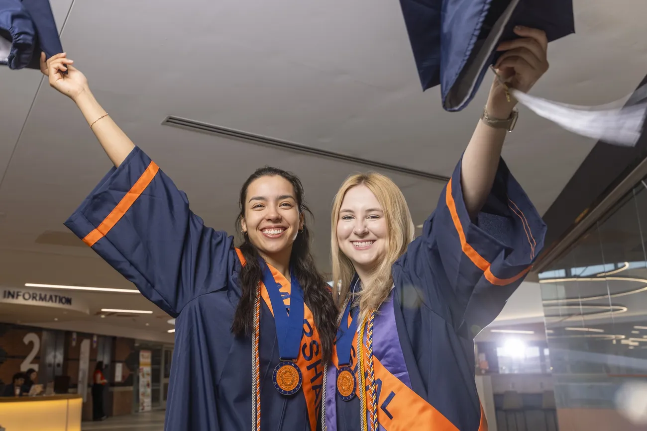 Student Marshals Ailed Torres Espinosa and Anna Meehan standing side by side holding up their graduation caps and smiling.