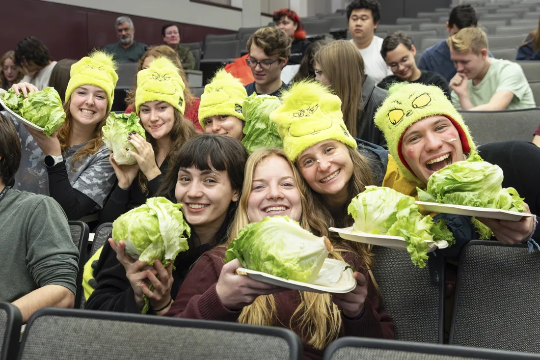 Students wearing Grinch-themed winter hats smiling with plates of lettuce heads.