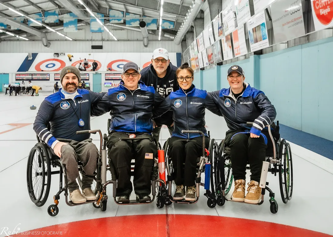 Katie Verderber smiling and posing on an ice rink with her teammates for Team USA's wheelchair curling team for the 2026 Winter Paralympic Games.