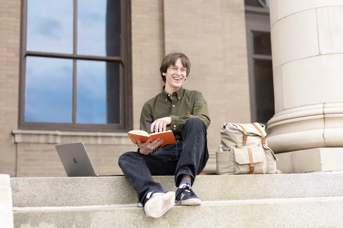 Rider Sulikowski '26 sitting outside on the steps of Carnegie Library holding a notebook and smiling.