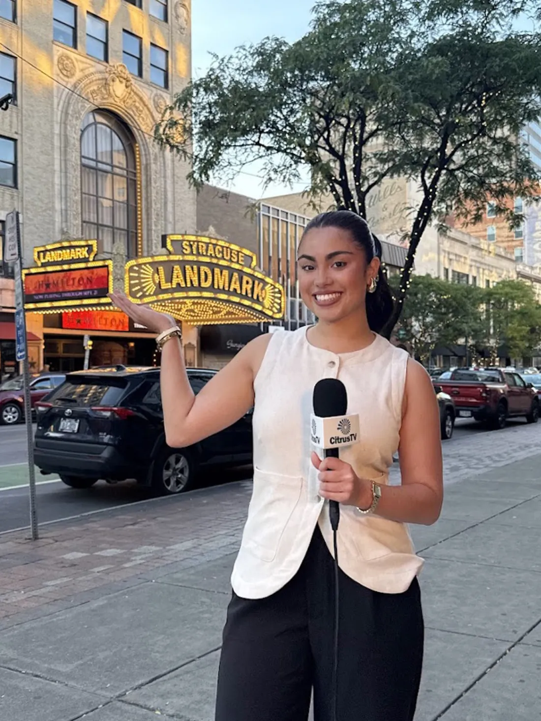 Junior Isabella Galan standing outside of Syracuse's Landmark Theatre holding a microphone for a CitrusTV broadcast.