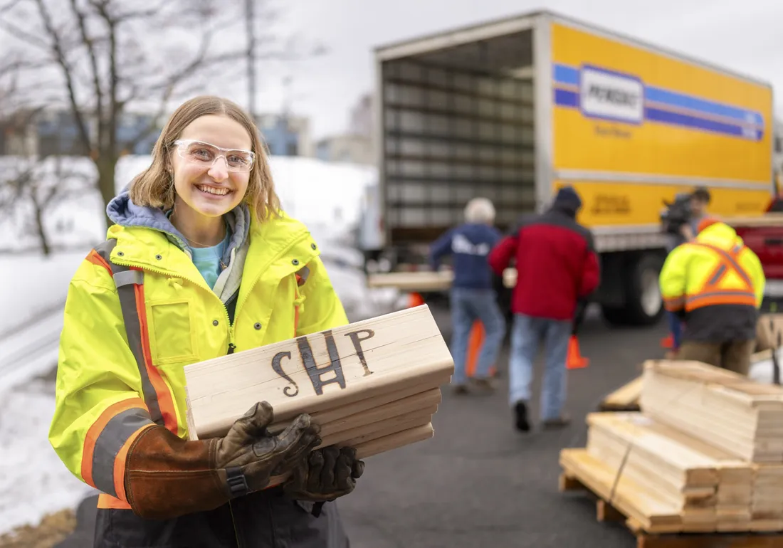 Claire Ceccoli '25 smiling and holding a stack of wood as part of a bed building event.