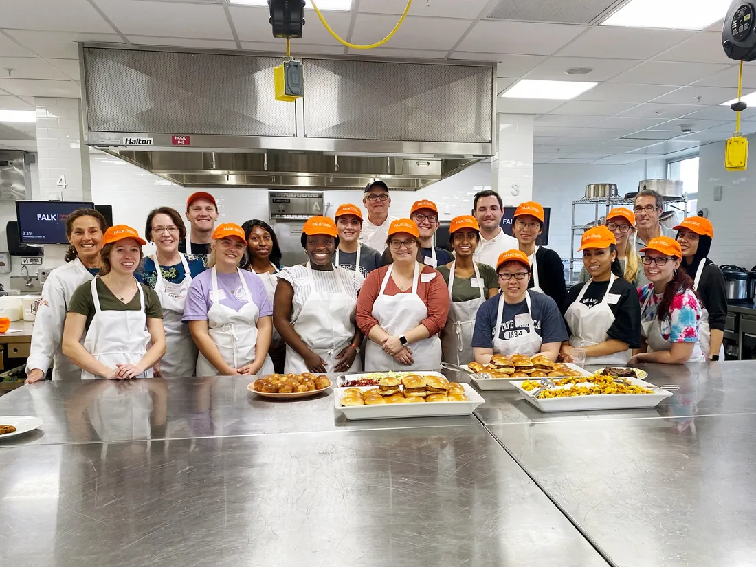 Group of people wearing apron standing in a commercial kitchen behind a table with dishes of food on it.