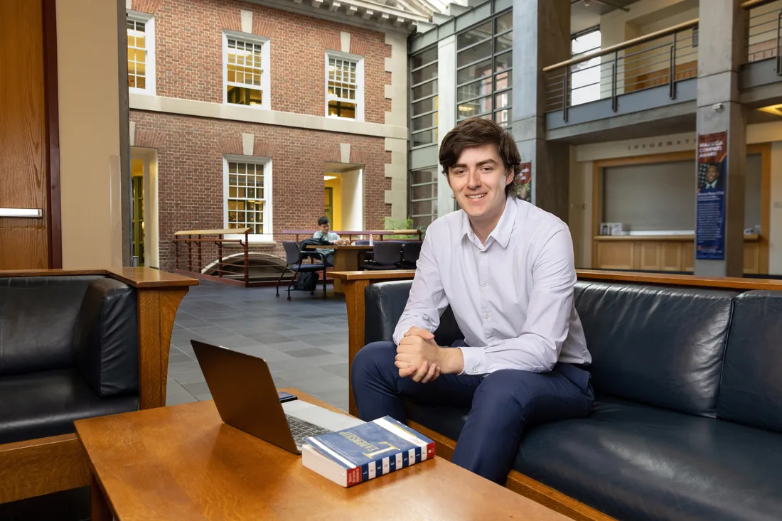 Portrait of David Bruen sitting on a couch with his laptop and a book, smiling with his hands clasped in front/