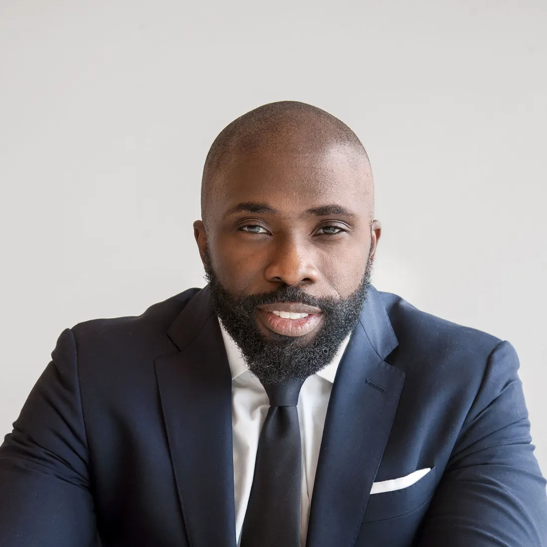 Leke Osinubi sitting in front of a white wall in a navy suit and smiling.