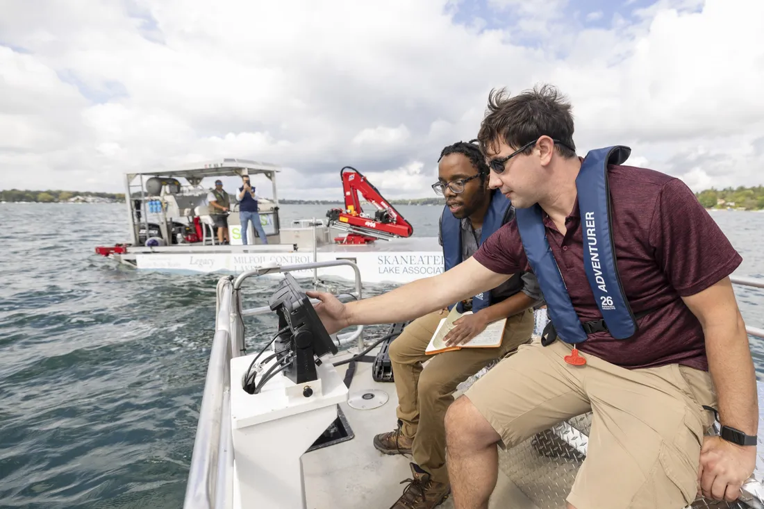 Two students sitting on a boat on Skaneateles Lake looking at a tablet screen monitoring algal blooms