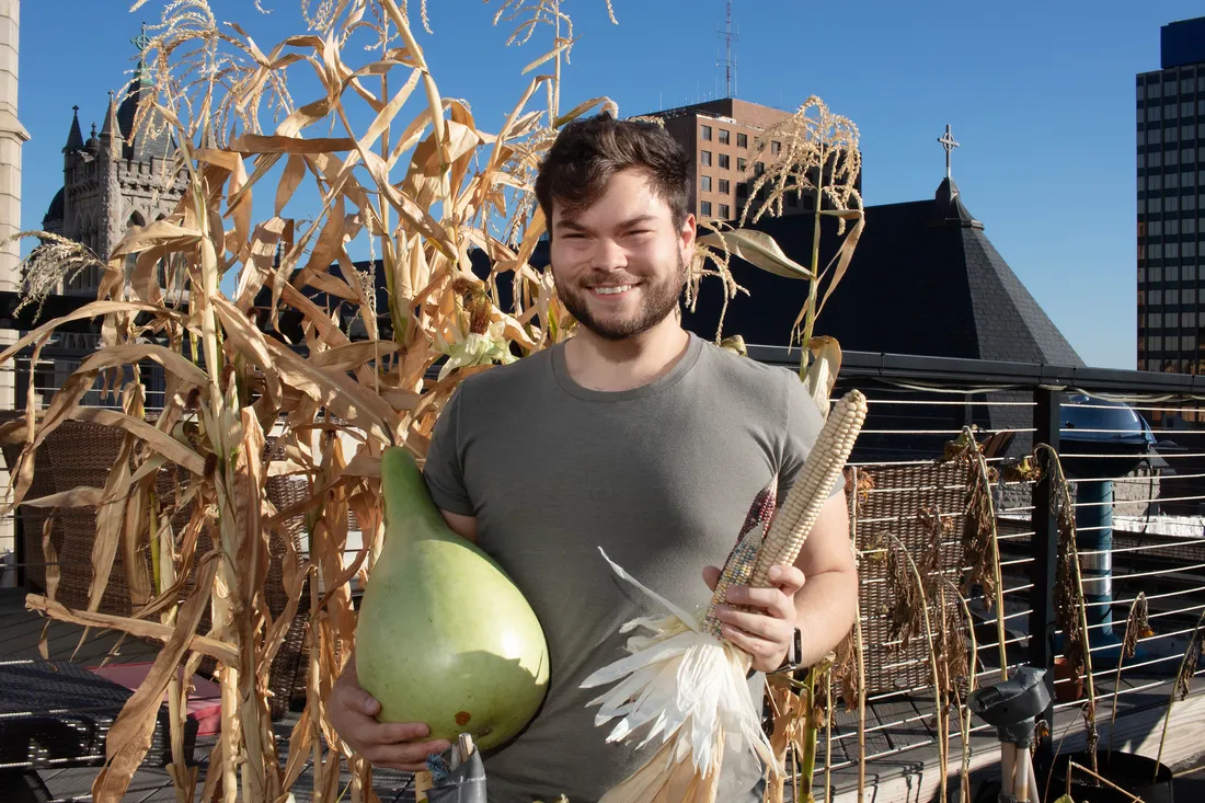Portrait of Ethan Tyo on his rooftop garden, surrounded by stalks and holding squash and corn, smiling.