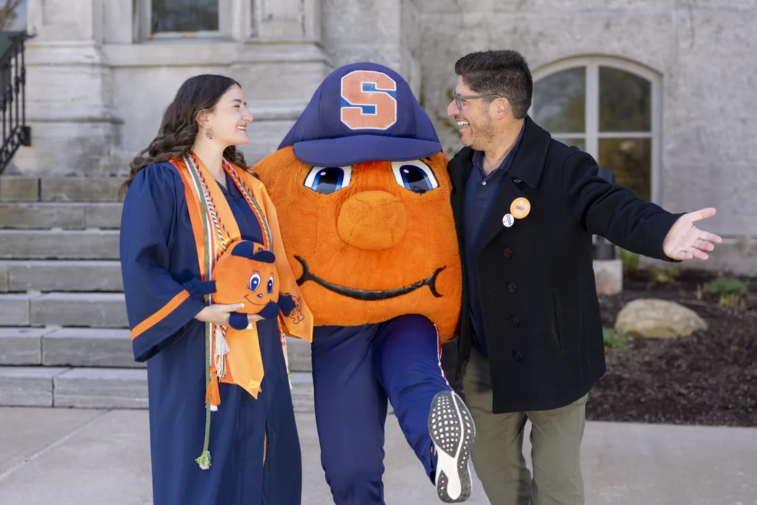 Sarah Jacoby, dressed in graduation regalia and holding an Otto the Orange plush toy, smiling and standing side by side with Otto and Mitch Messinger.