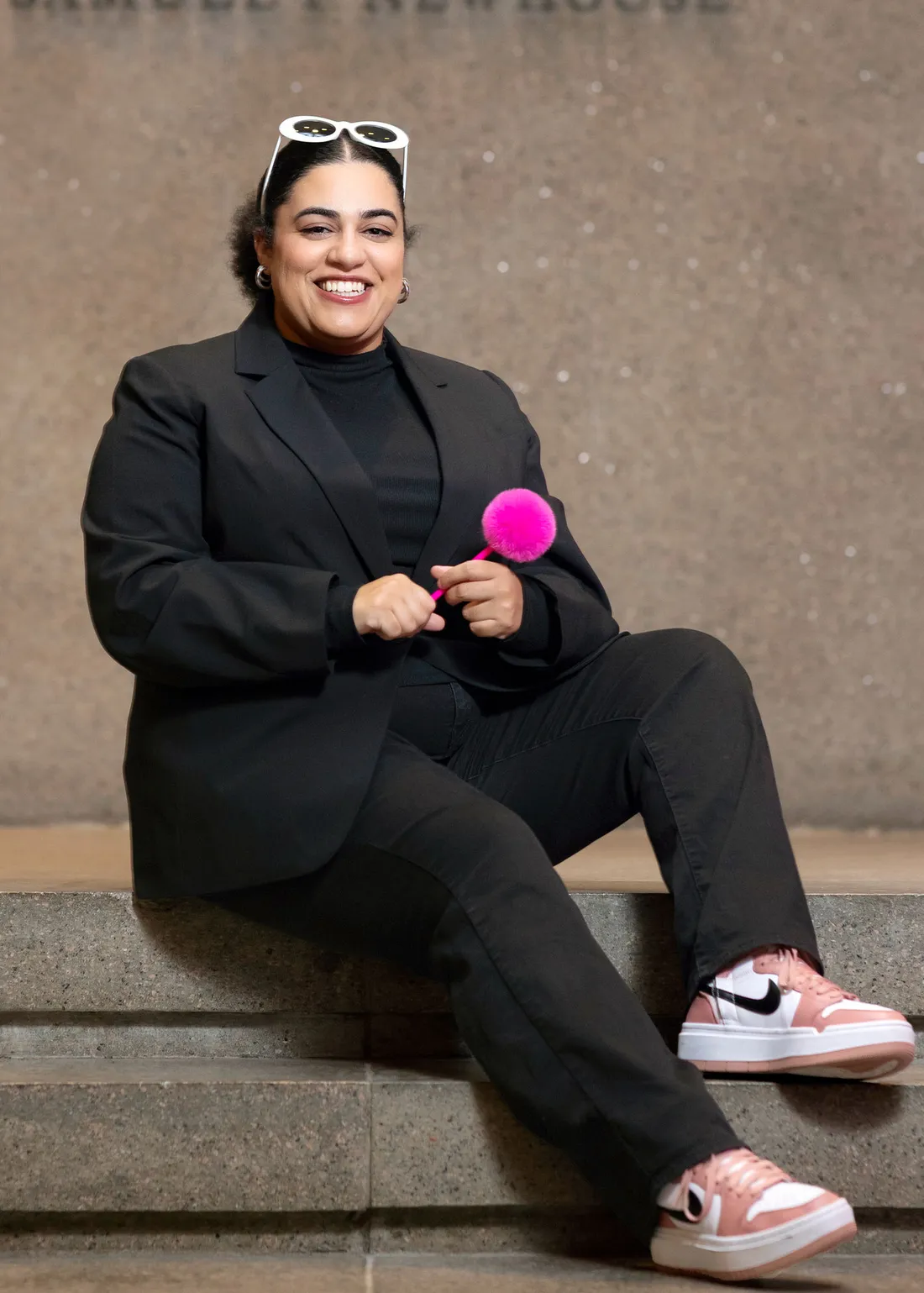 Carmela Boykin holding a pink pen and sitting on steps inside the Newhouse School of Public Communication.