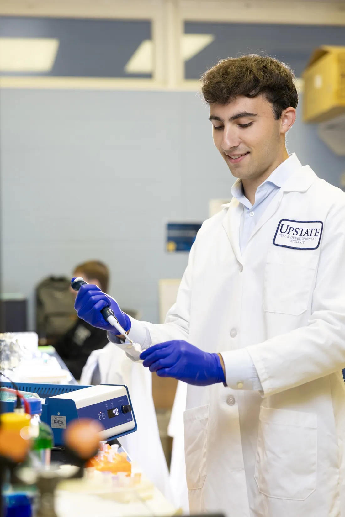 Beckam Polis ’28 wearing a lab coat and gloves and using a pipette in a laboratory.