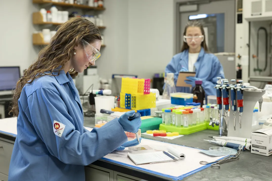 Two people sitting at a lab desk using pipettes and taking notes.