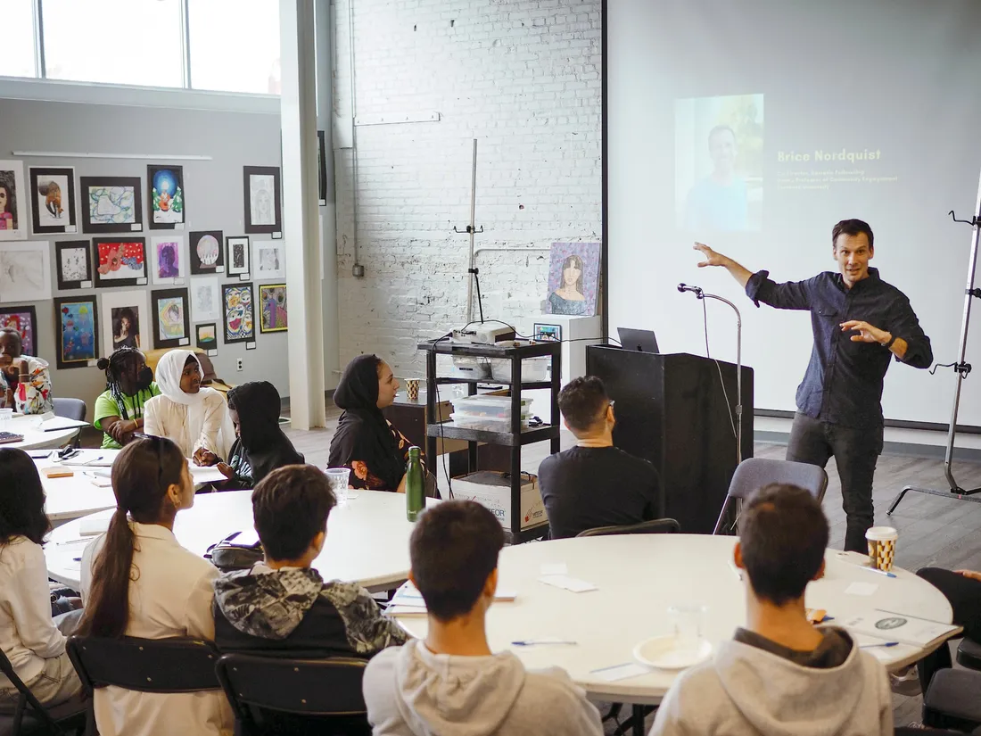 Brice Nordquist talking at the front of a classroom in front of seated students, artwork on the wall behind him.