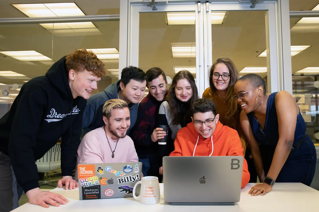 A group of people look over the shoulder of a student working on a laptop.