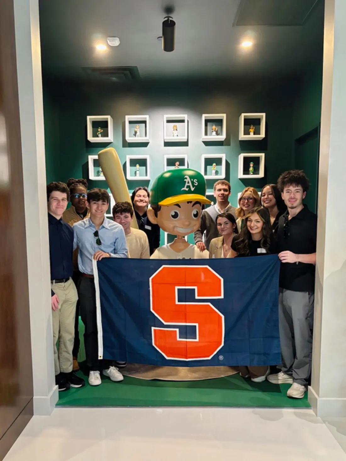 Students in the Las Vegas spring immersion program standing inside the Las Vegas A's Ballpark Experience Center holding a Syracuse University flag.