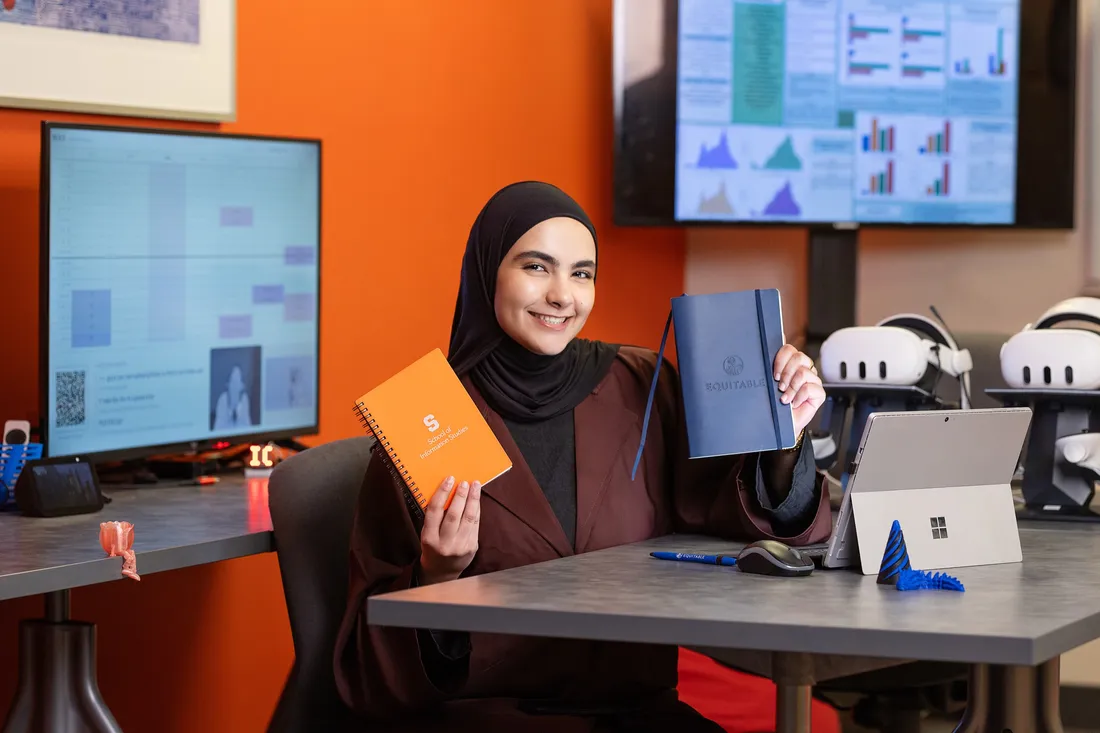 Bushra Khazaleh '26 sitting in the Nexis student technology lab holding two notebooks and smiling.