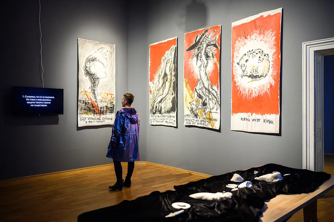 Woman stands in front of panels of multimedia art in gallery hanging on gray wall.