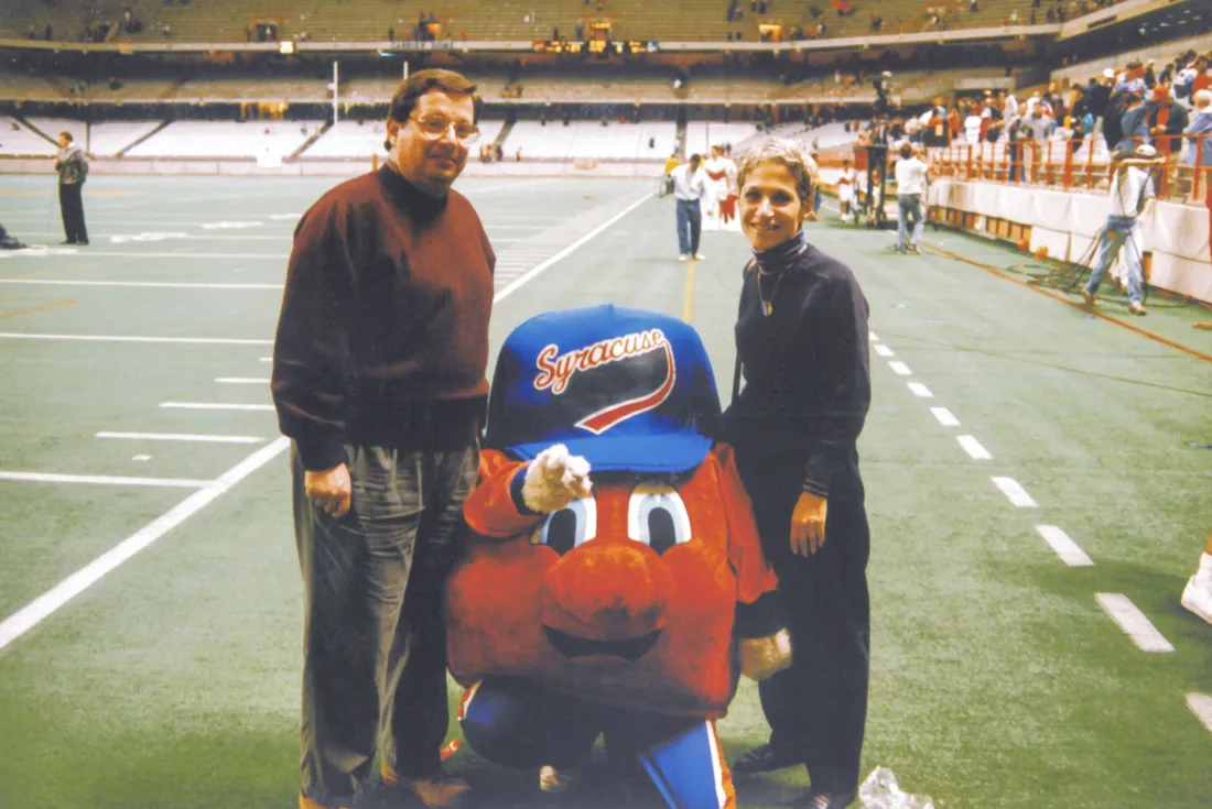 Mitch Messinger dressed in the original Otto the Orange mascot suit, posing with his parents on the sidelines of a game.