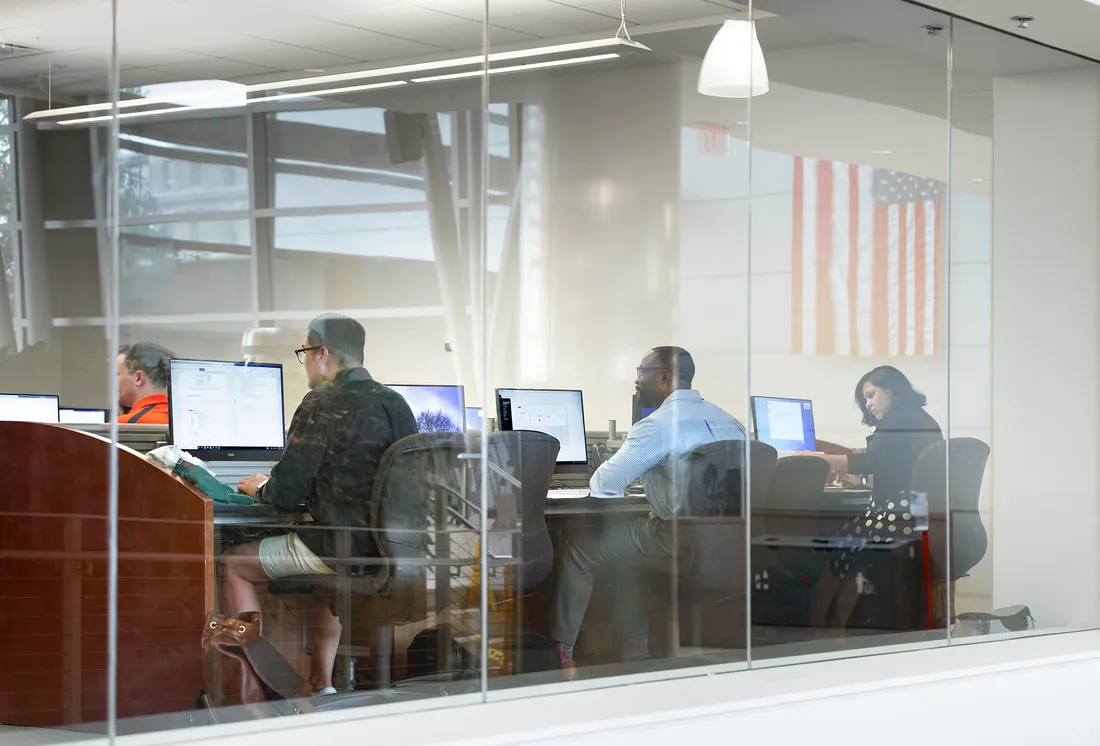 Landscape photo of students working on their computers behind the glass wall of a classroom.