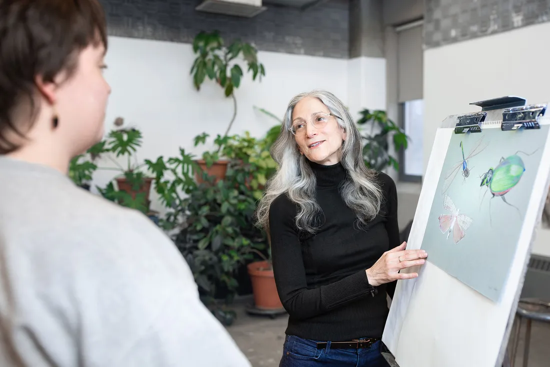 A woman stands at a easel and provides feedback to a student whose work is on display.