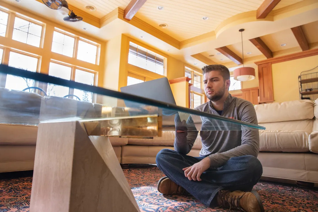 Edward Furcinito sitting on the floor of his house, with his laptop open on a glass coffee table.