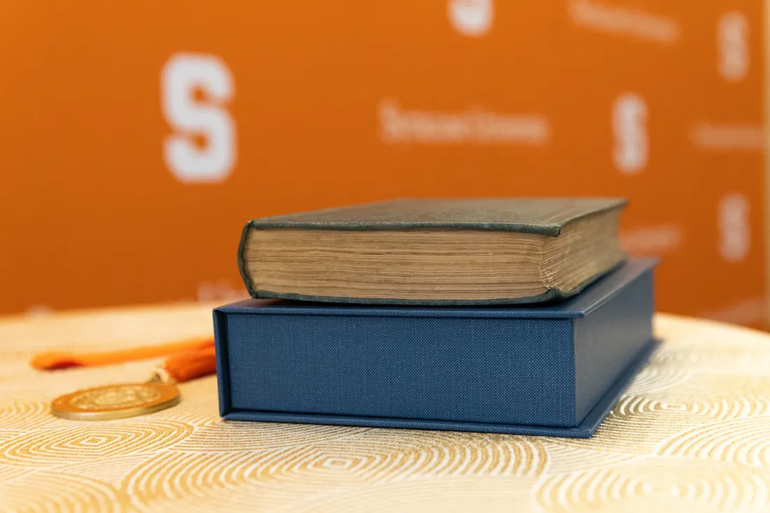 Textbooks are stacked on a desk with Syracuse University background.