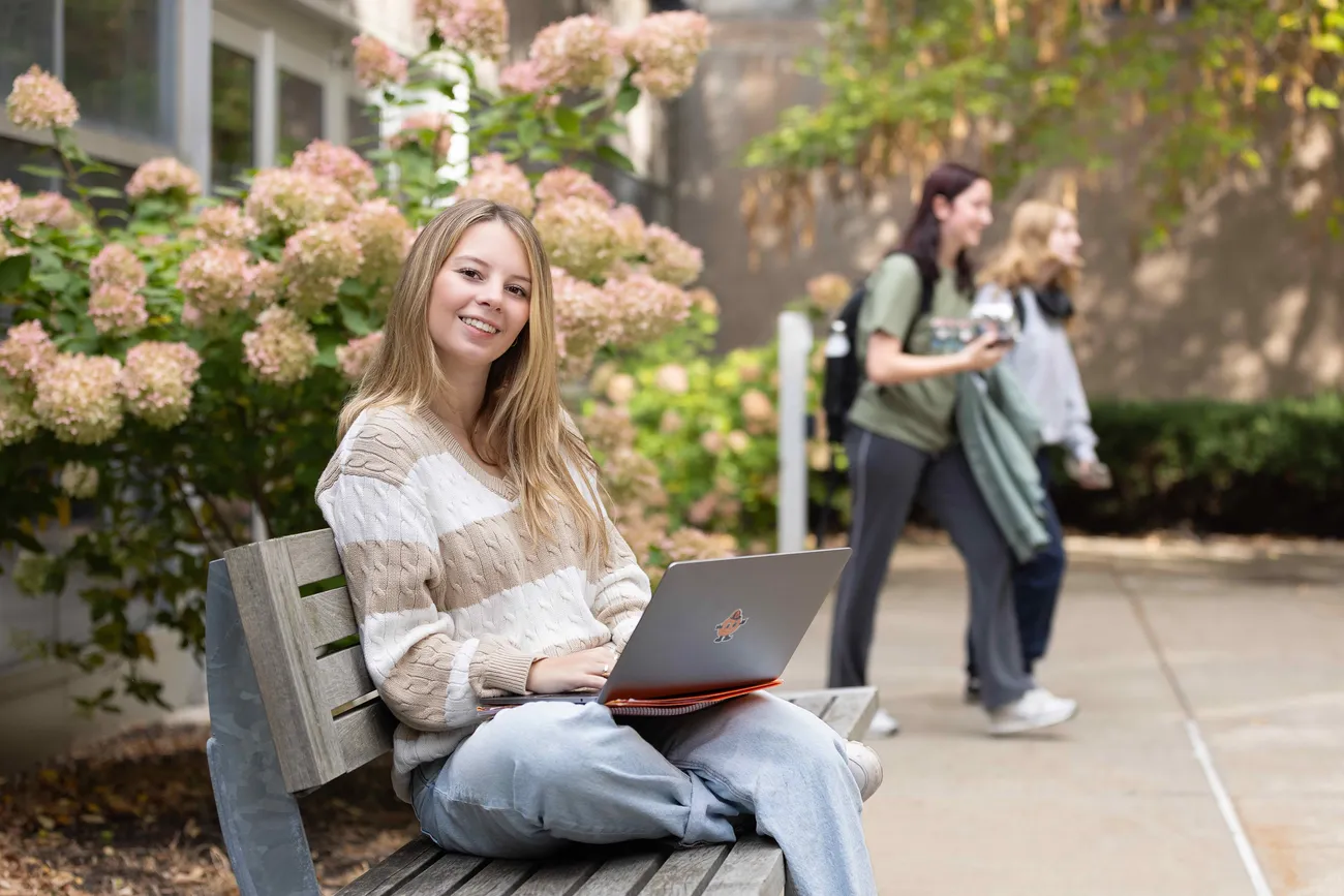 A student sitting outside smiling on a computer.