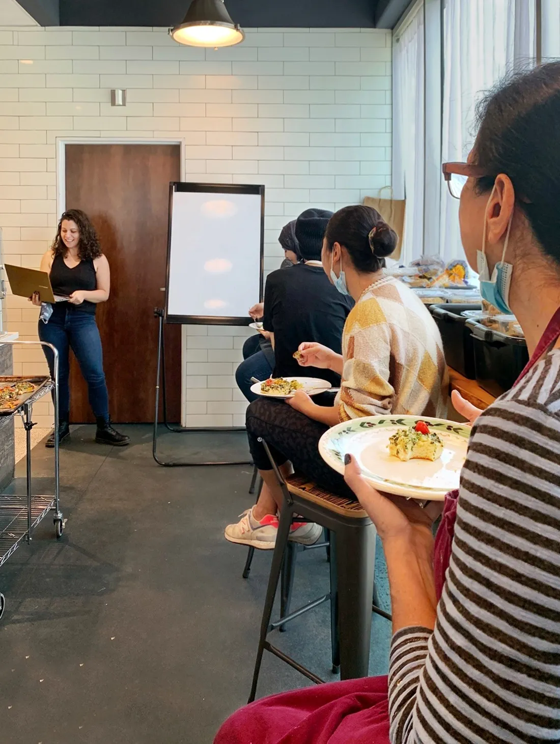 Cara Santino standing at the front of a room next to a whiteboard, speaking to a group of people sitting at small tables with plates of food.