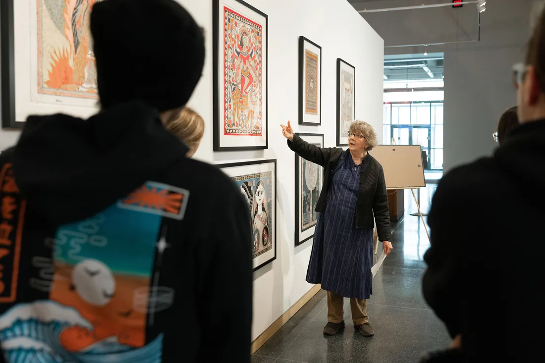 Woman in art gallery points to paintings while students look on.