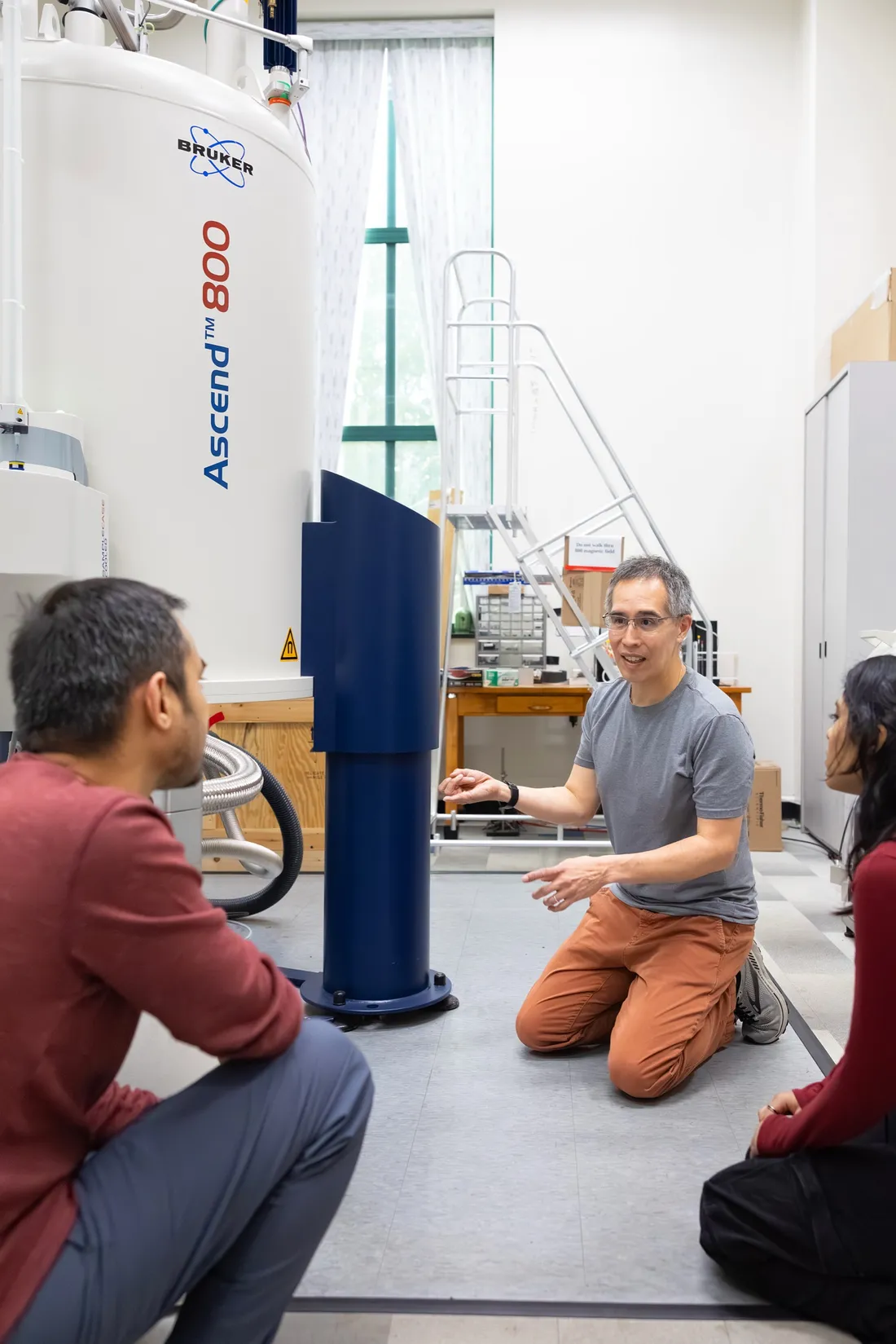 Professor Carlos Castañeda sitting on the ground with two students showing them a nuclear magnetic resonance spectrometer.